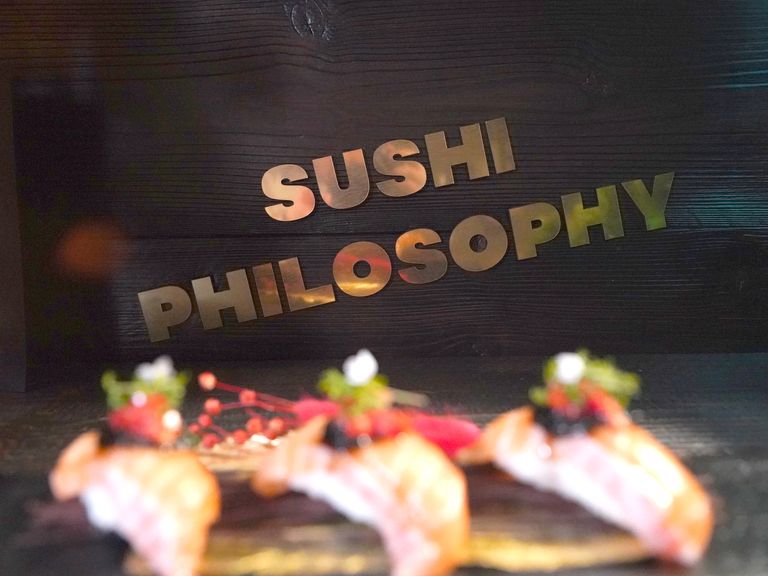 Schriftzug Sushi Philosophy aus poliertem Edelstahl vor dunkler Wand, im Vordergrund Sushi